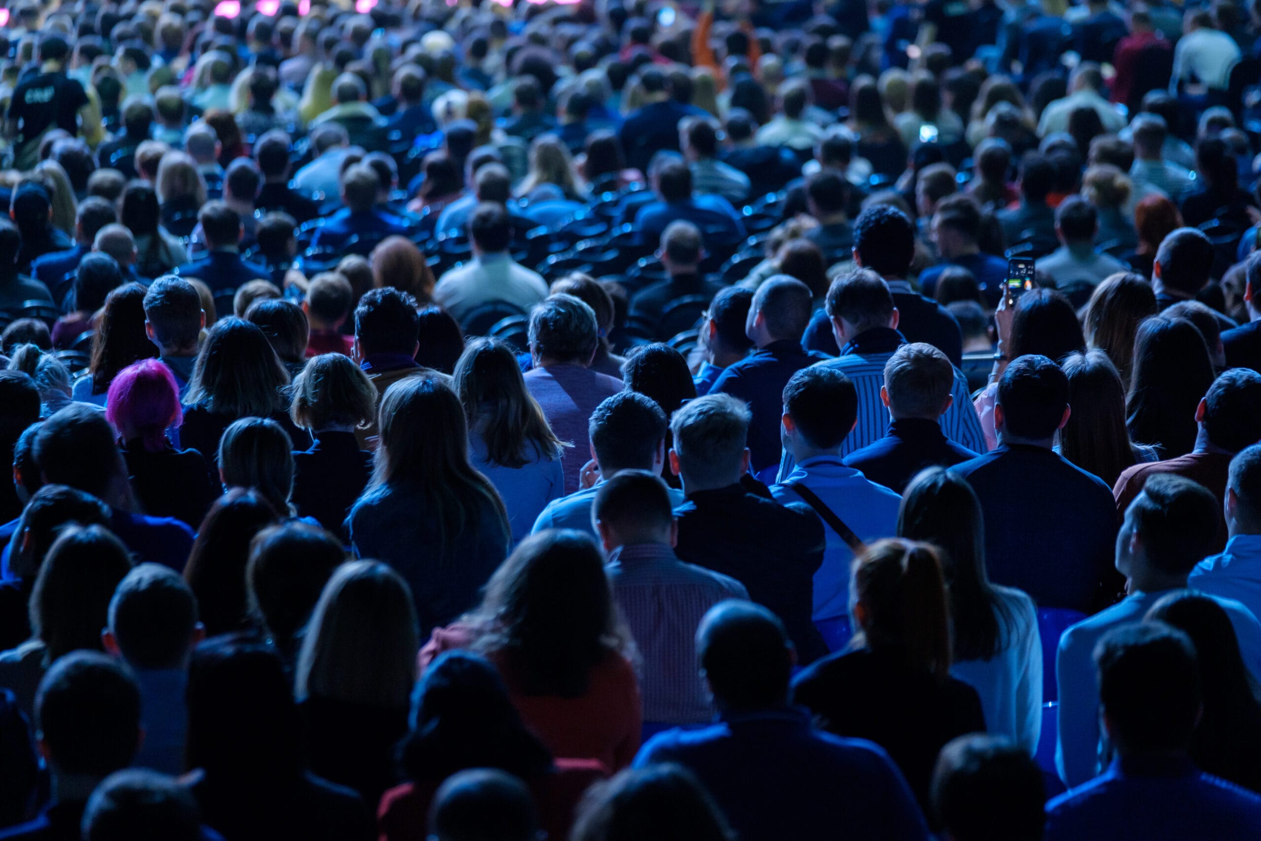 A photo of a crowd of people with backs turned towards us watching someone on stage. Photo has blue over tone.