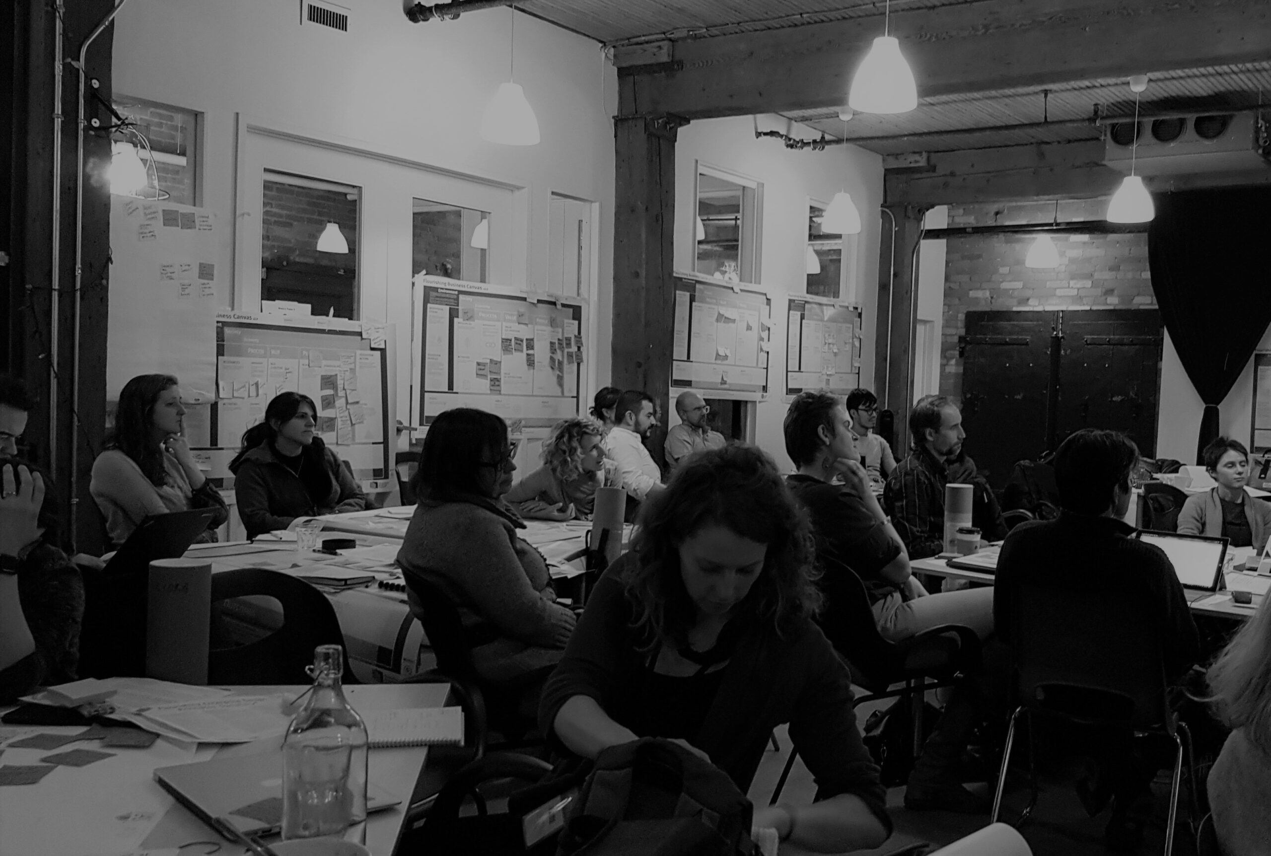 A black and white image of a group of about 20 people in a workshop. Posters with sticky notes on the surrounding walls